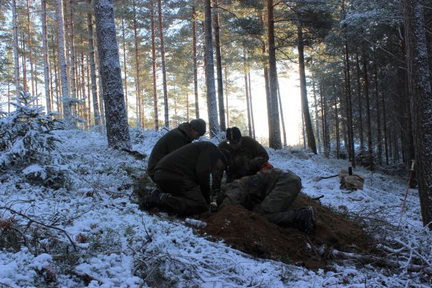 Useita metsästäjiä kerääntyneenä yhteen kaivamaan maata talvisessa metsässä. 