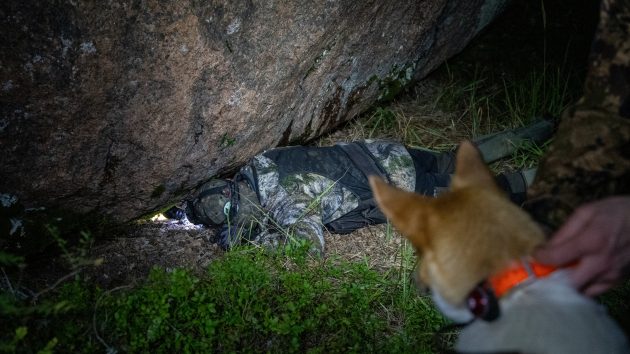 Jägaren ligger på marken och tittar under en stor sten. Det finns en hund i närheten.